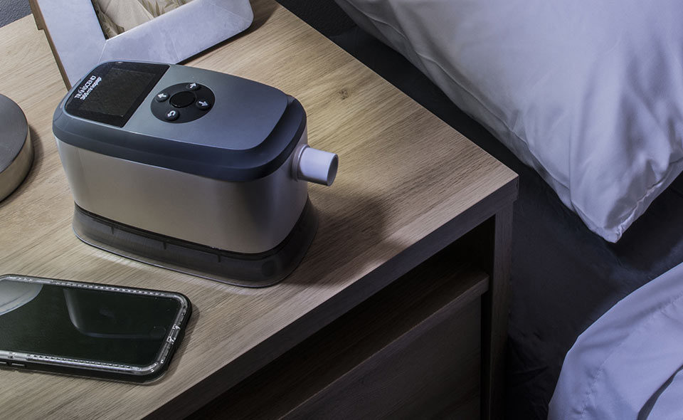 A CPAP machine sits on a wooden bedside table next to a smartphone, lamp, and a bed with white pillows and sheets.