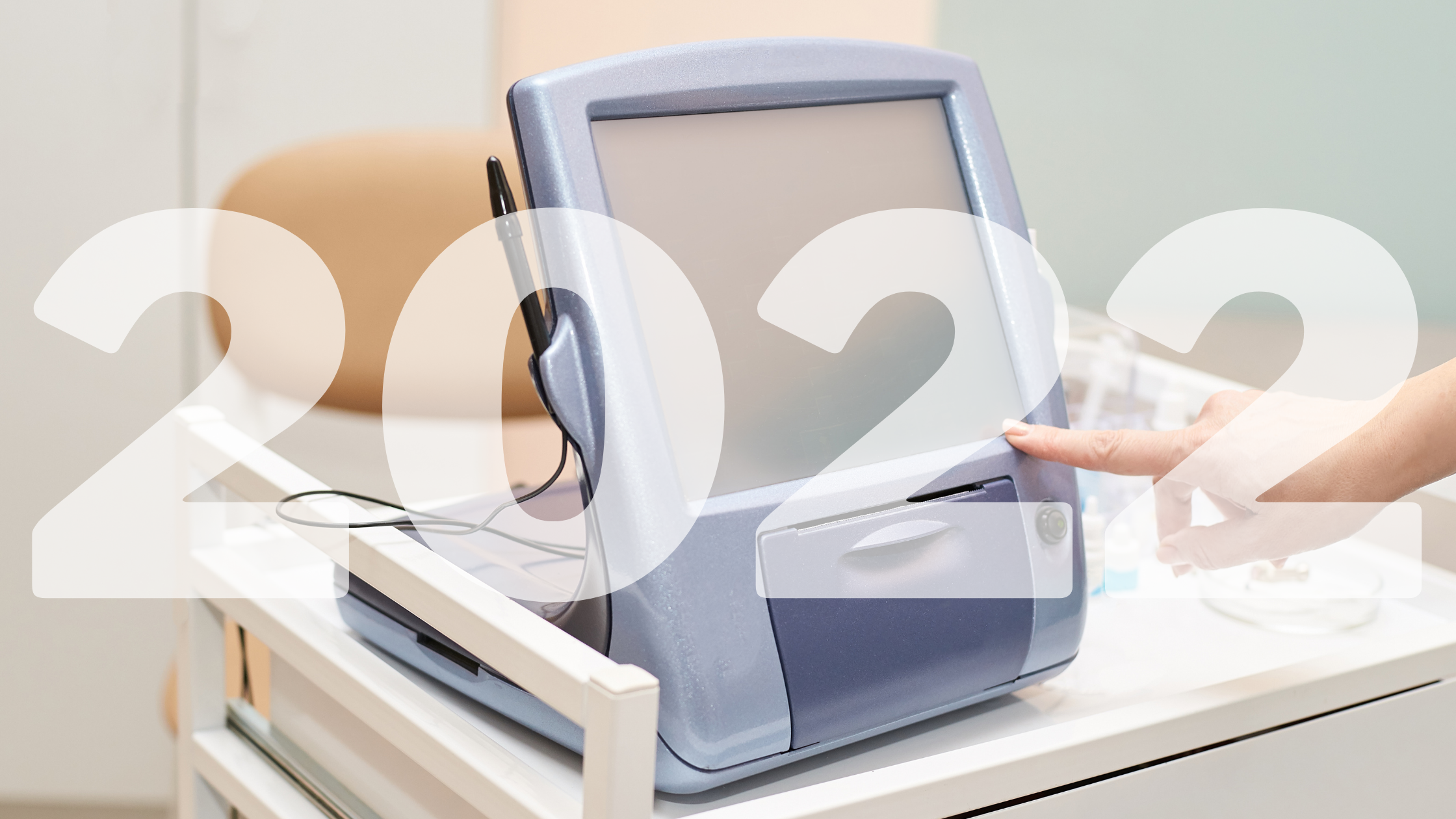 A person’s finger touches a touchscreen medical device on a cart. Large, semi-transparent numbers “2022” are overlaid across the image.
