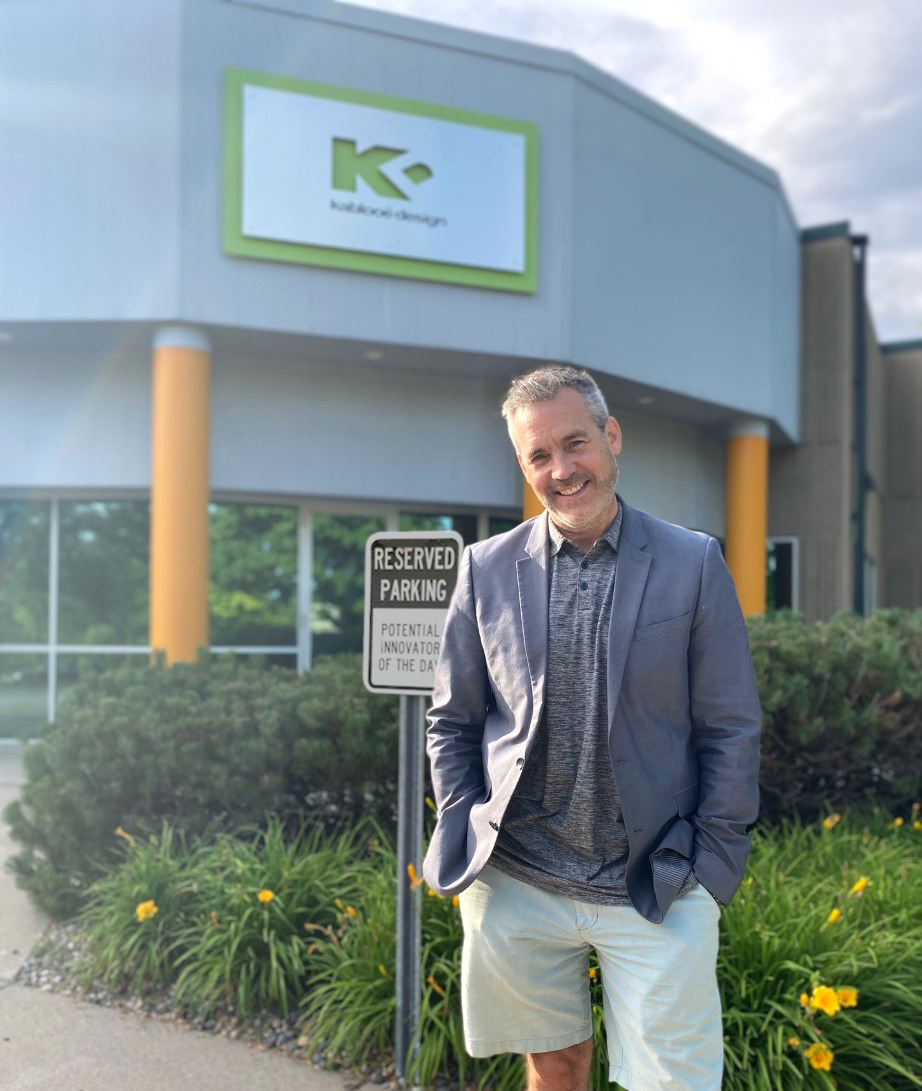 A man in a blazer and shorts stands smiling with hands in pockets in front of a building with a green "K" logo and a "Reserved Parking: Potential Innovator of the Day" sign. Yellow flowers and bushes are in the foreground.