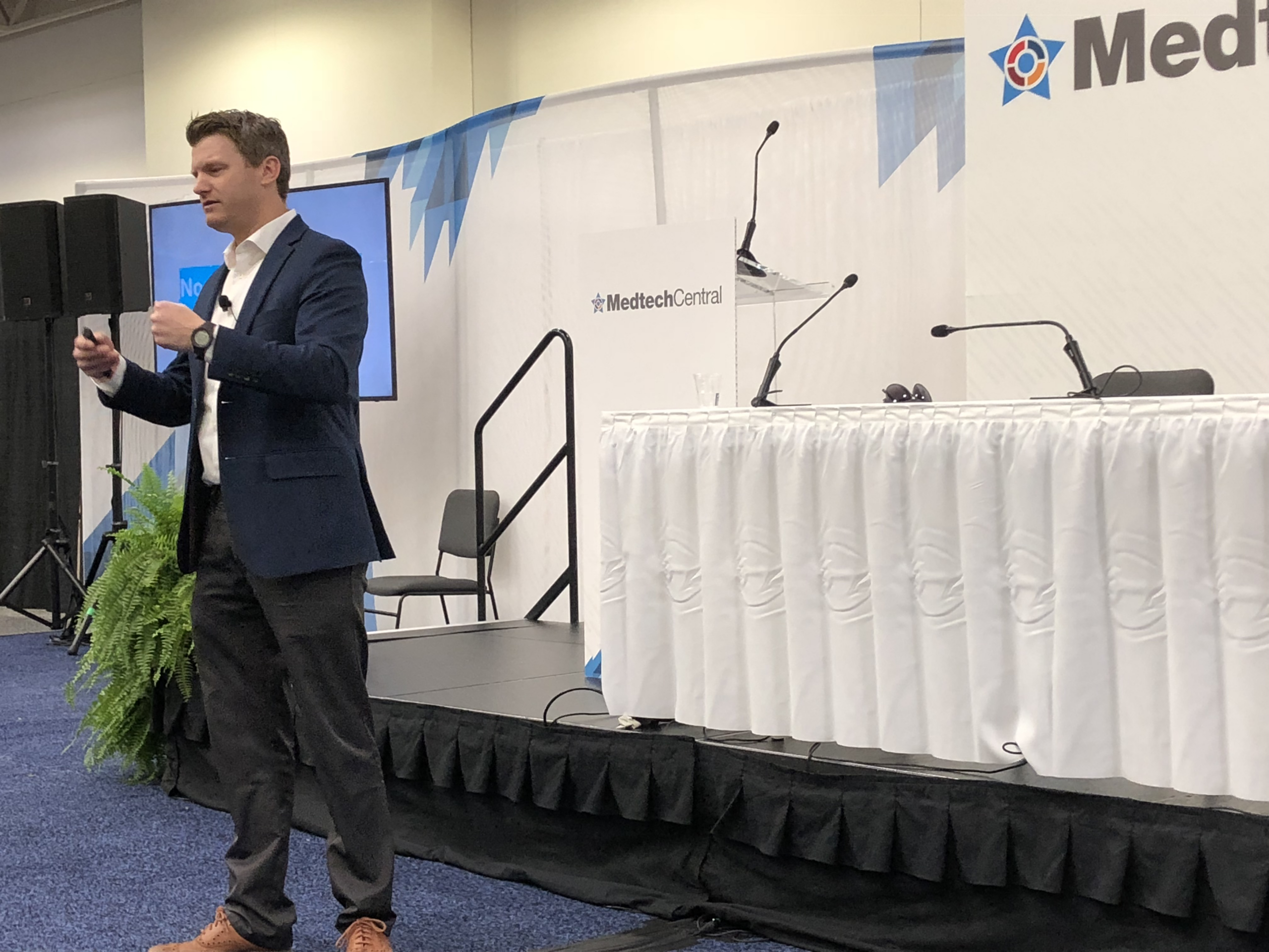 A man in a navy blazer speaks onstage at a conference, holding a microphone. Behind him is a table with white skirting, microphones, and a MedtechCentral banner. A monitor and potted plant are also visible.