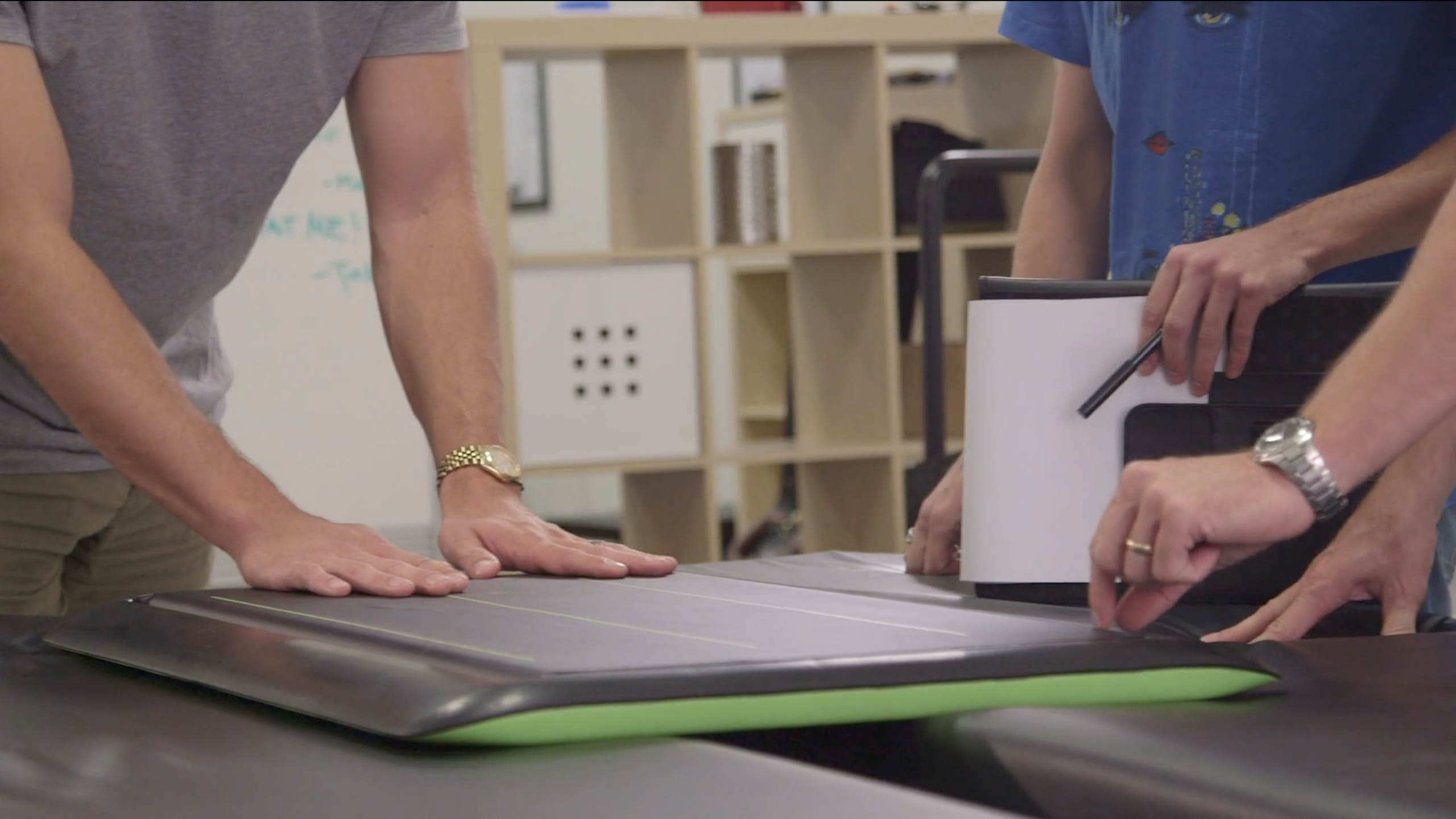 Two people stand at a table, examining a large, flat, rectangular object with a green edge. One person holds a tablet with a stylus, while the other places hands on the object. Shelves and a whiteboard are visible in the background.