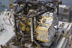 A close-up of a spacecraft component in a laboratory, covered in foil insulation with an arrow pointing to a specific metallic section on the right side. Equipment and cables are visible in the background.