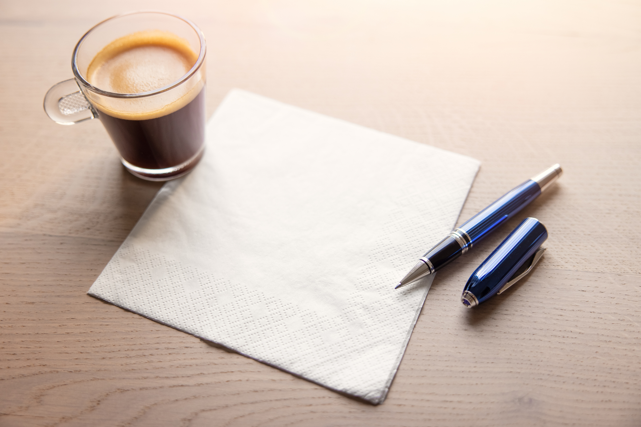 A cup of coffee sits on a wooden table next to a blue pen, its cap removed, resting on a white paper napkin.
