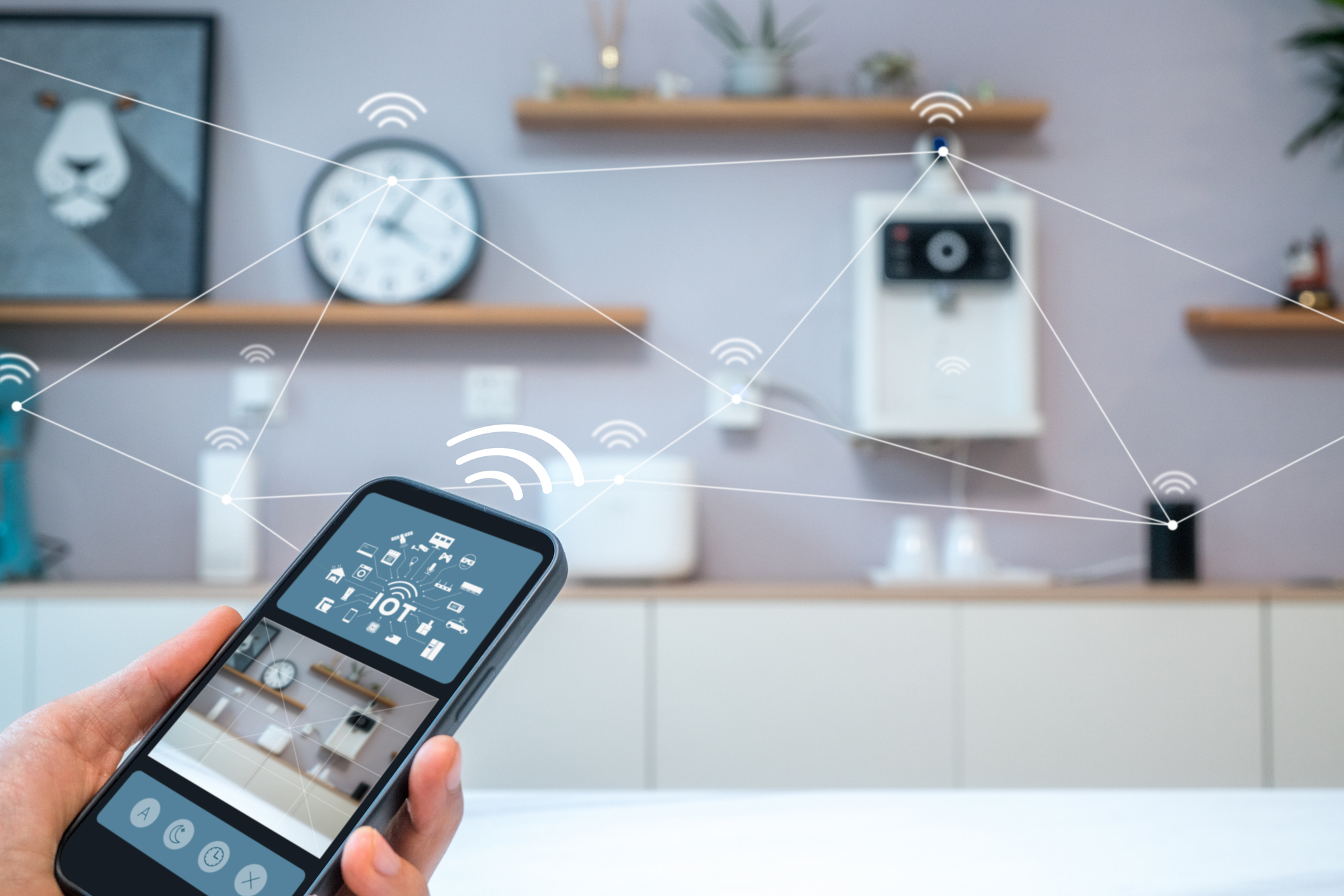 A hand holds a smartphone displaying a smart home control app, with wireless connection symbols linking devices in a modern, furnished room in the background.