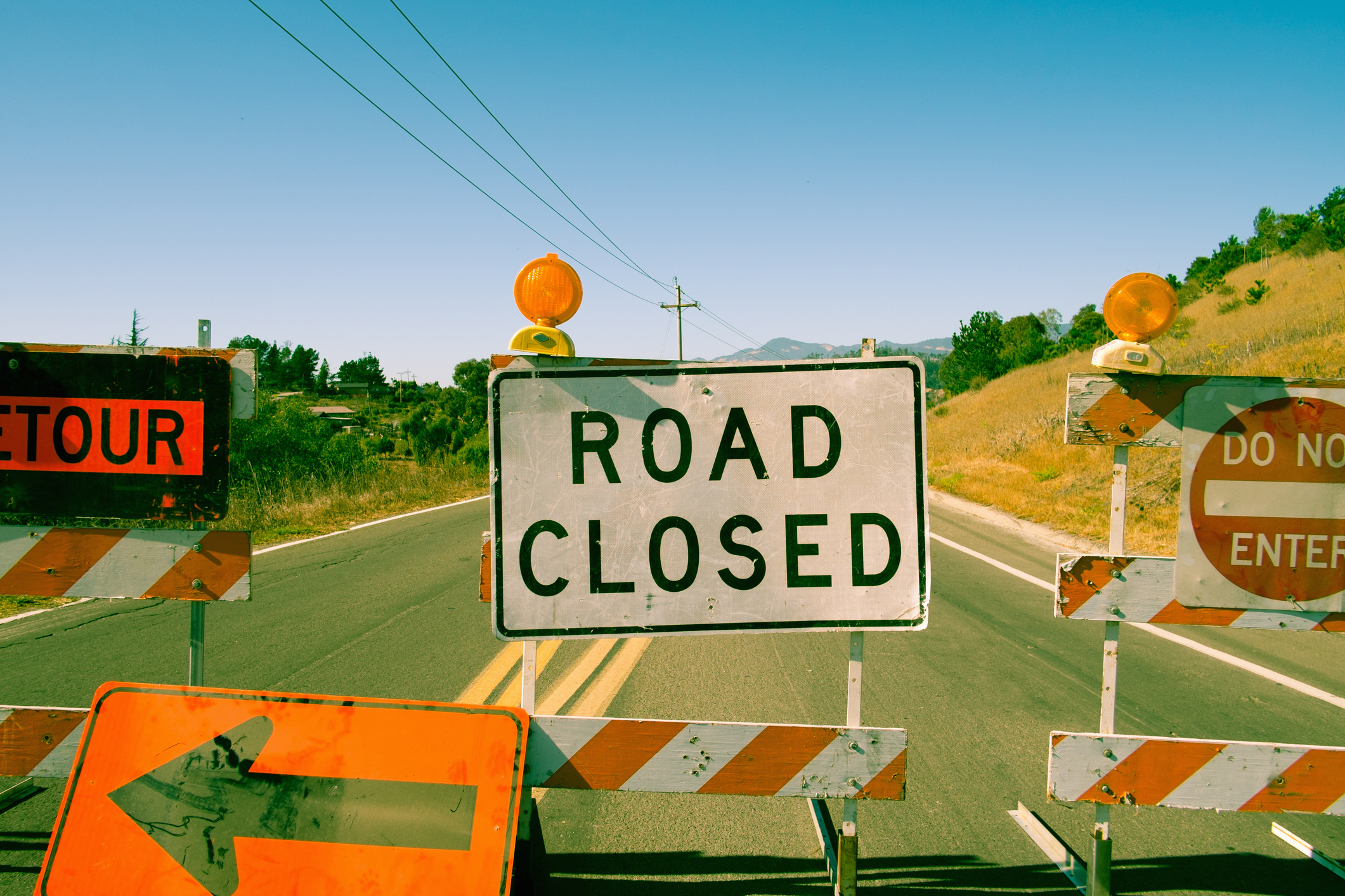 A "Road Closed" sign blocks a rural road, flanked by barricades with orange lights. Additional signs indicate a detour to the left and warn not to enter. Grass and trees line the roadside under a clear blue sky.
