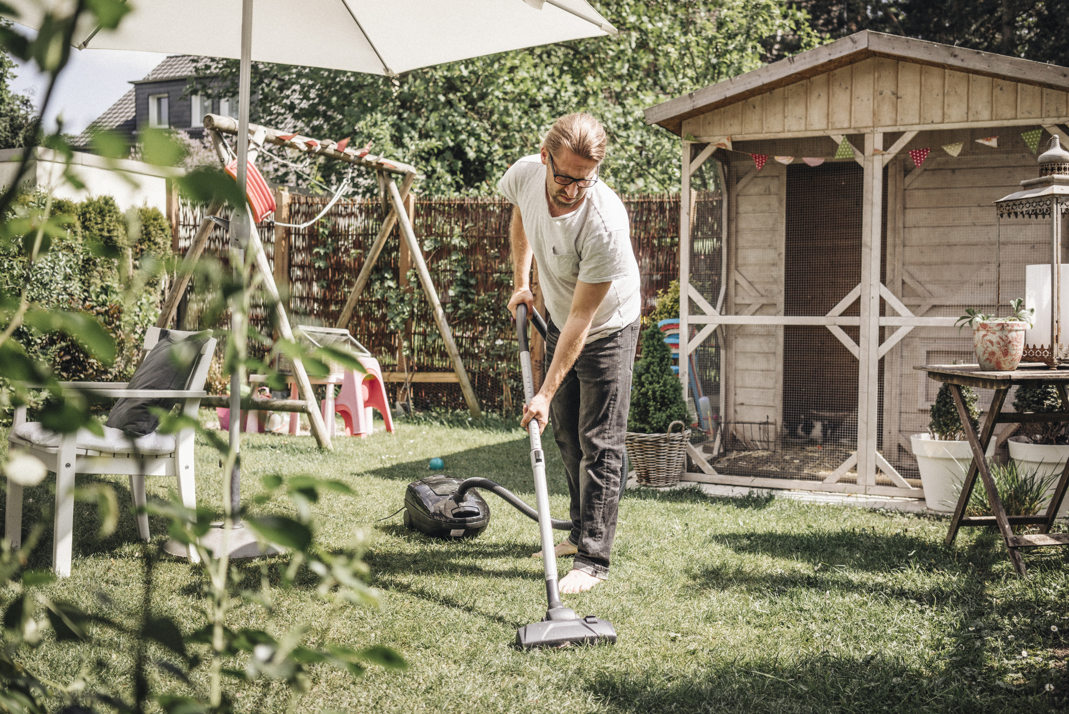 A man with glasses vacuums the grass in a sunny backyard, surrounded by garden furniture, a small wooden shed, a swing set, and plants.