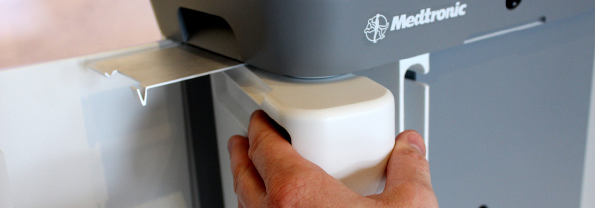 A hand holds a white container while inserting it into a gray Medtronic medical device, with part of a display and tray visible.
