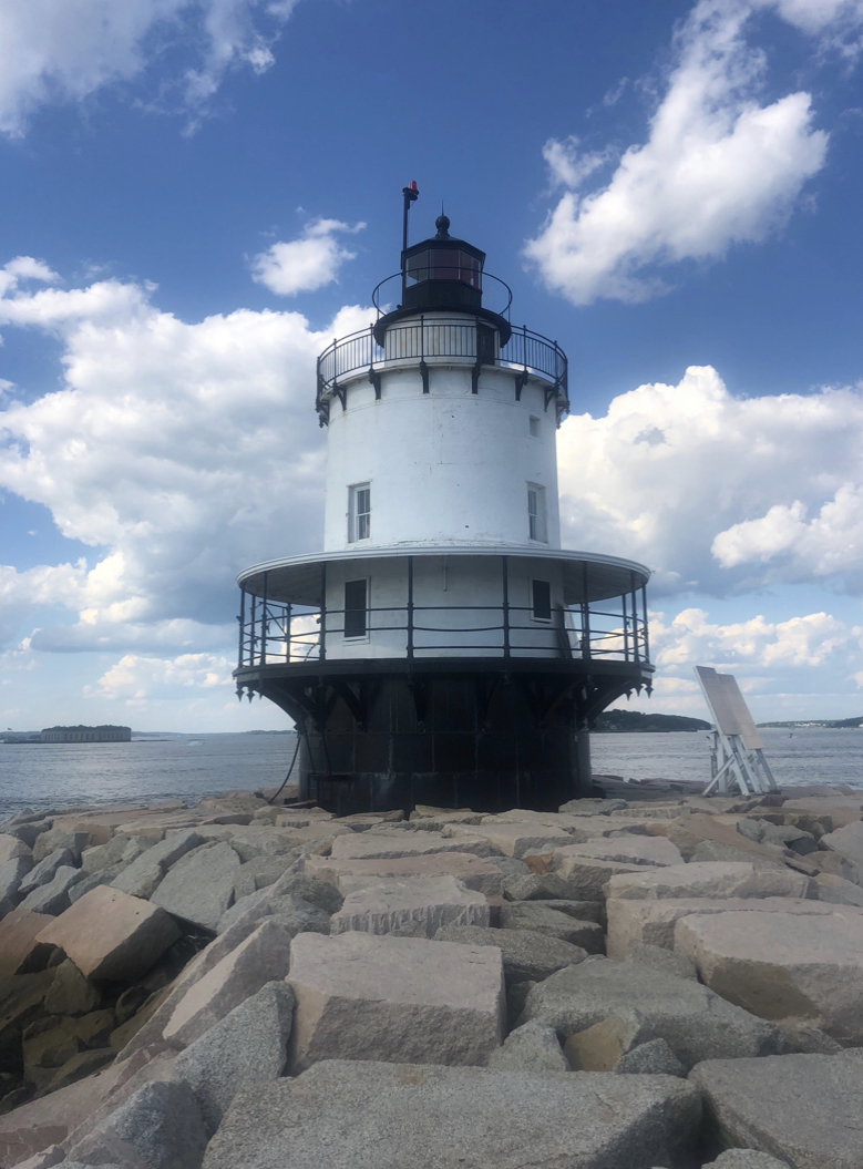 A white cylindrical lighthouse with a black base stands on large rocks by the water under a partly cloudy sky.