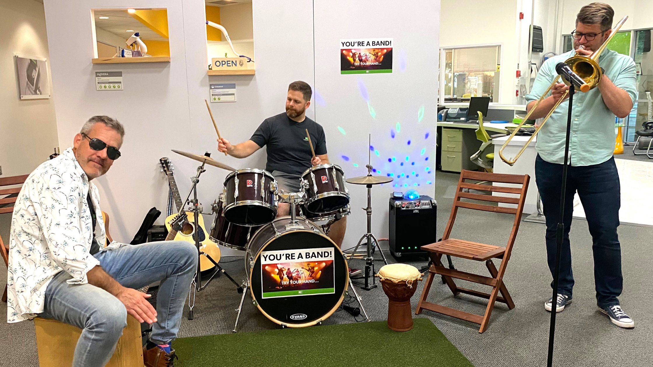 Three men play instruments indoors: one on a cajón drum, one on a drum set, and one on a trombone. They are surrounded by chairs and instruments, with a sign reading "YOU'RE A BAND!" in the background.