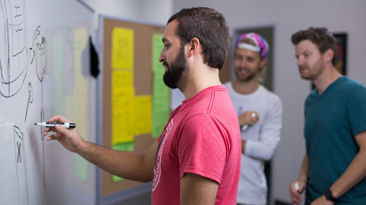 A man in a red shirt draws on a whiteboard while two other men stand nearby, watching and smiling. There are colorful sticky notes on a corkboard in the background.