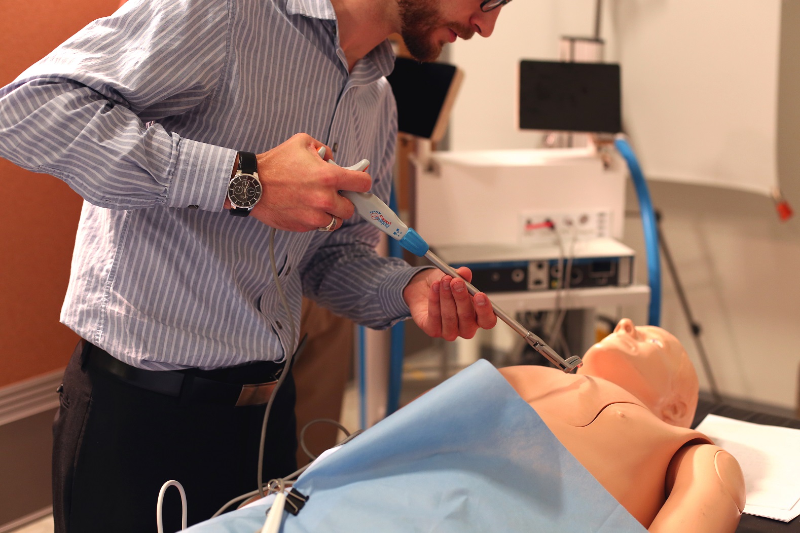 A person in a striped shirt practices a medical procedure on a medical training mannequin, using a syringe-like instrument, in a clinical training environment.