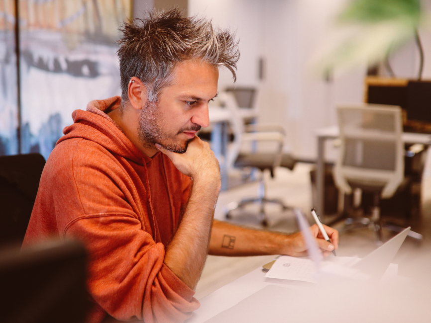 A man with gray hair wearing an orange hoodie sits at a desk in a modern office, looking thoughtful as he writes notes on paper with a pen.