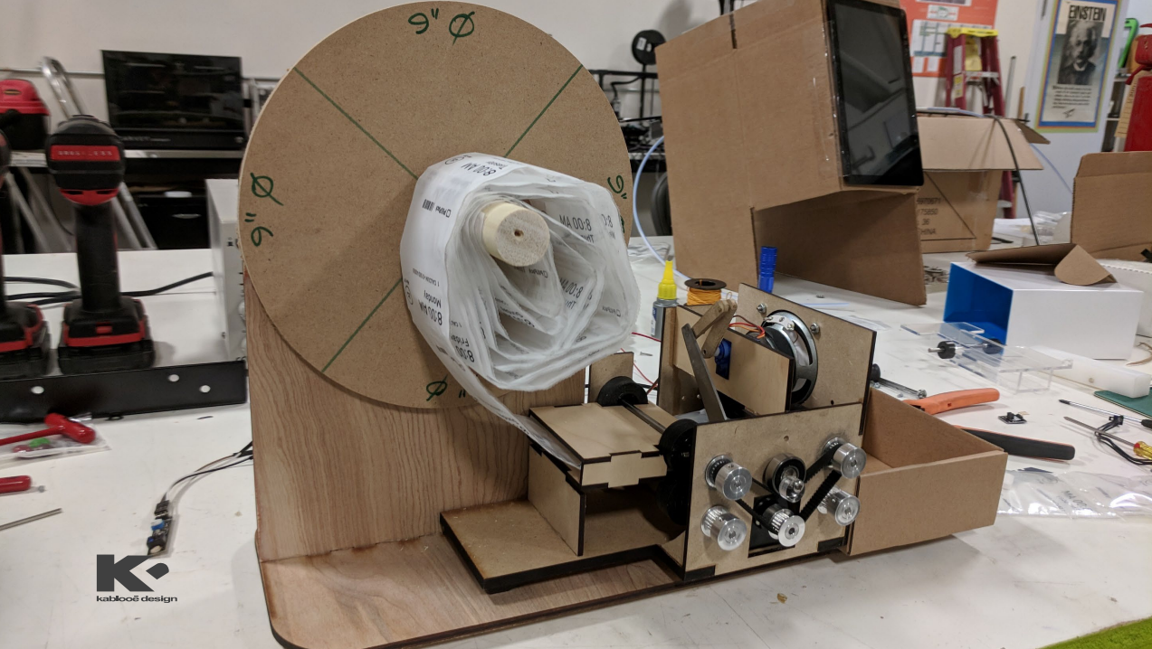 A homemade mechanical device made of wood and cardboard holds a large spool of plastic tubing, with gears, wheels, and electronic components arranged on a cluttered workbench. Tools and other workshop items are in the background.
