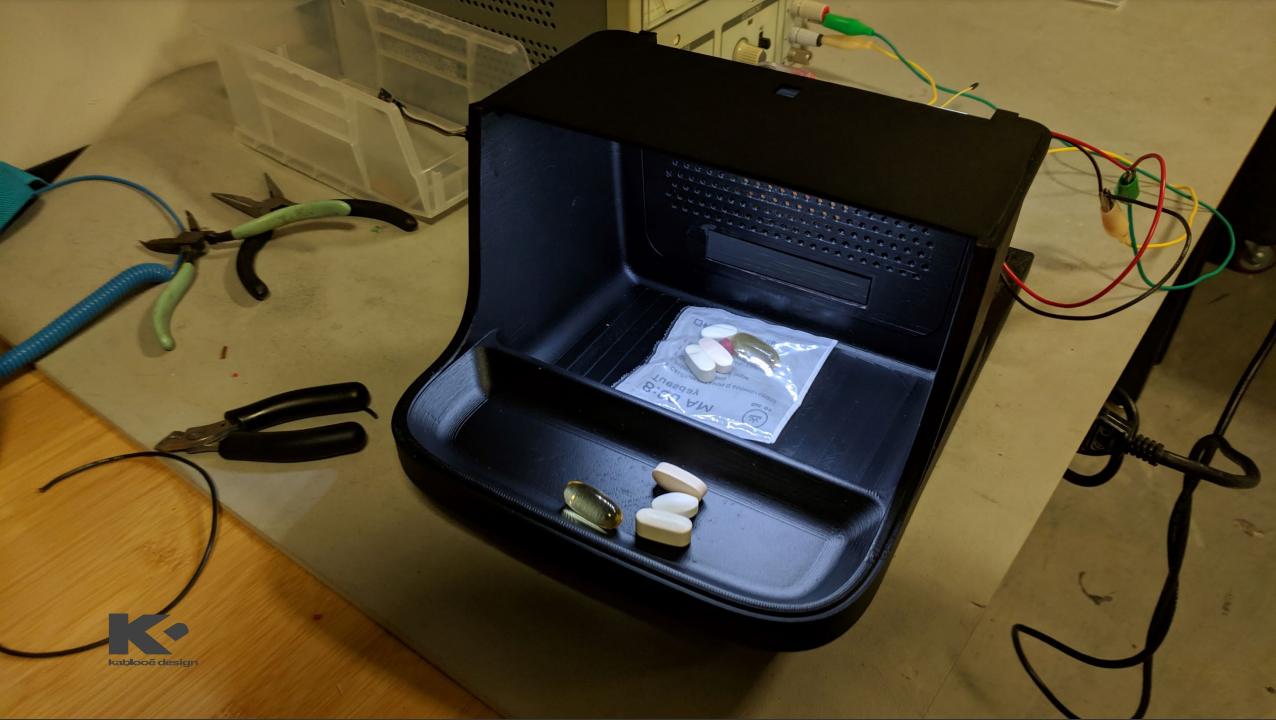 A black device sits on a workbench, illuminated inside and containing various pills, some loose and some in a plastic bag. Tools, wires, and containers are scattered around the workspace.