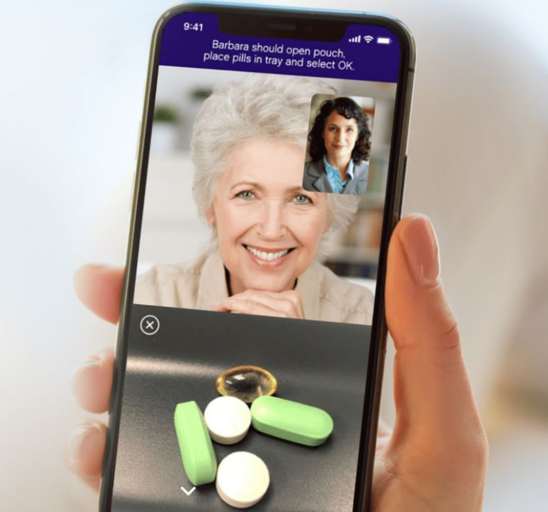 A hand holds a smartphone showing a video call between two women, with the screen also displaying a tray of pills including green tablets and white round pills.