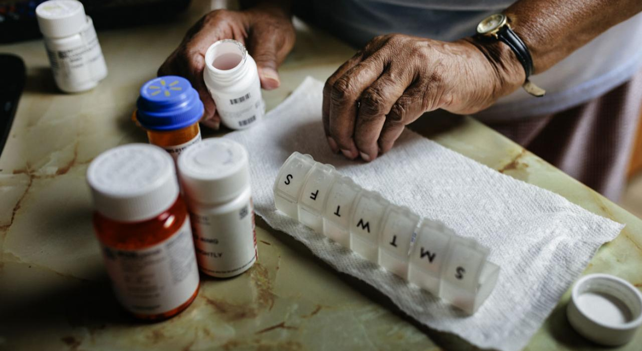 An elderly person’s hands organize pills into a weekly pill organizer labeled with days. Several prescription pill bottles are on a table beside them.