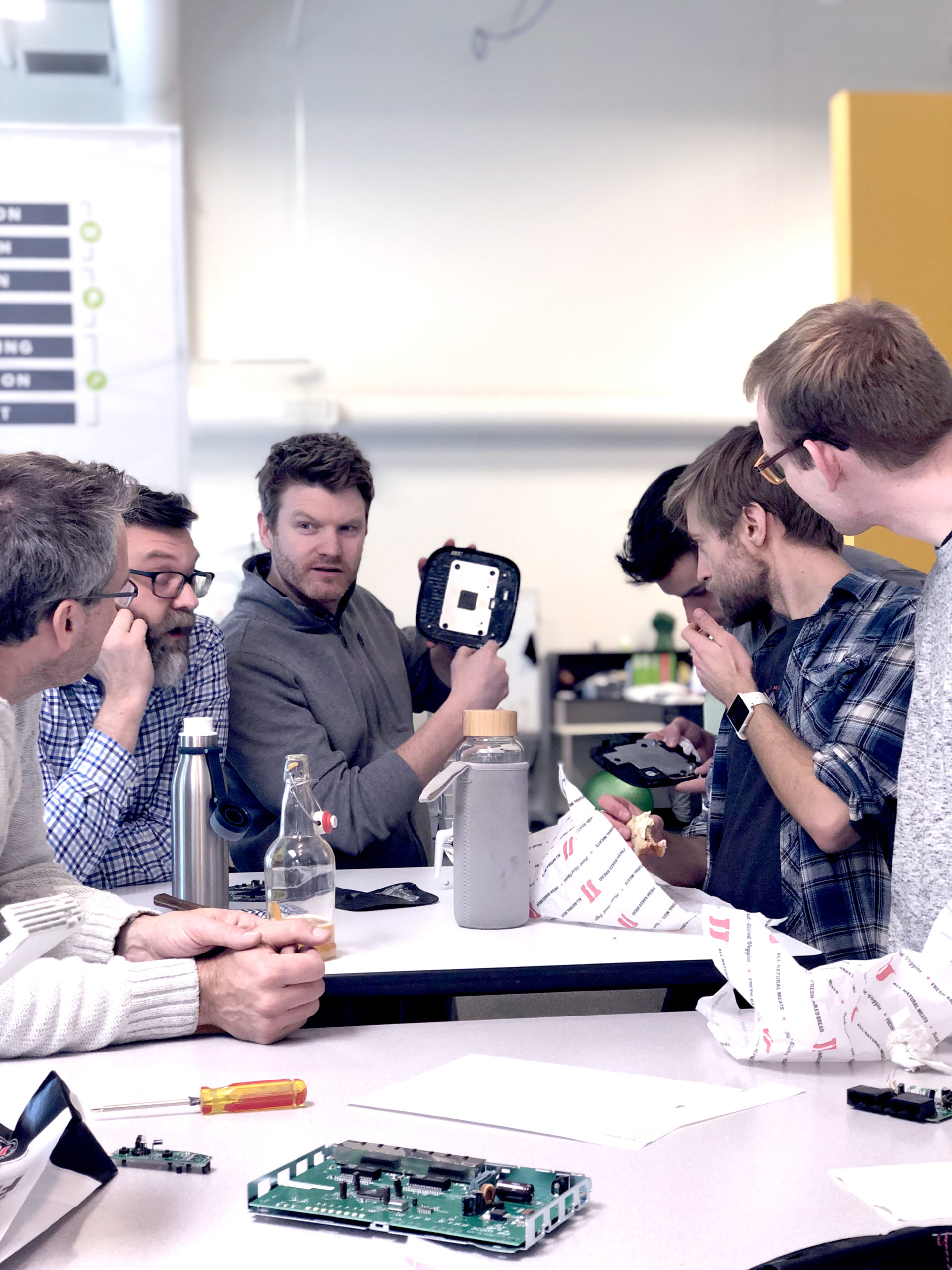 Six people sit around a table covered with electronic components and tools. One person holds up a device while others listen and examine parts. They appear to be discussing or troubleshooting the device in a collaborative setting.