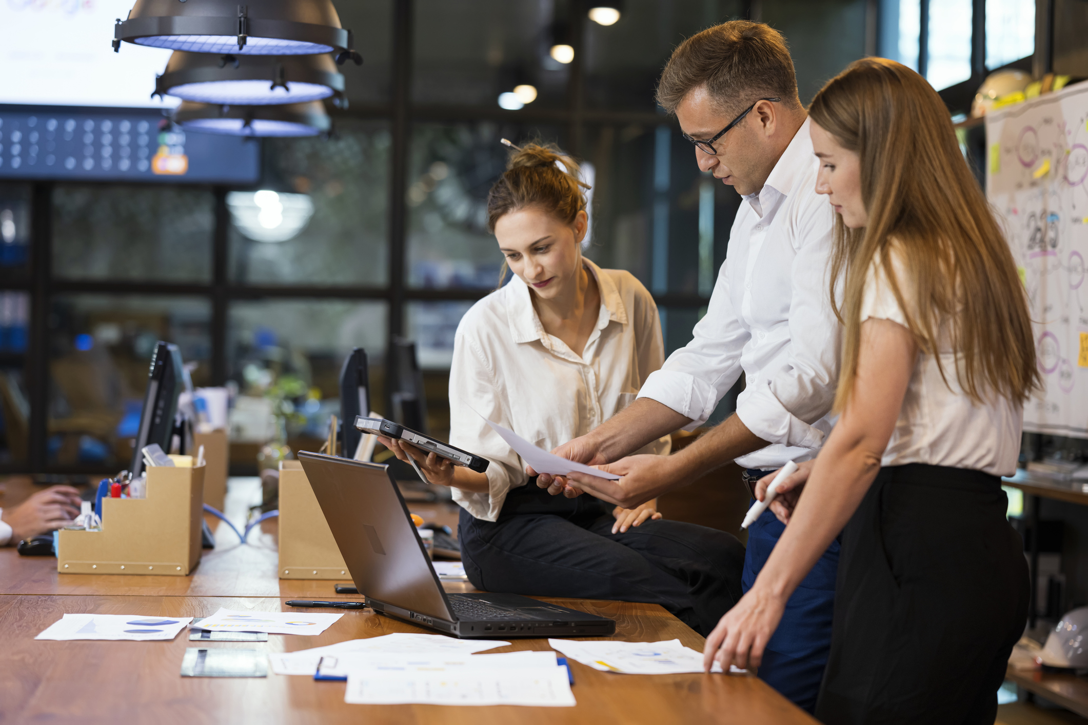 Three people collaborate at a wooden table in a modern office. They review documents and use a laptop, with papers and office supplies scattered around. One person sits on the table while the others stand.