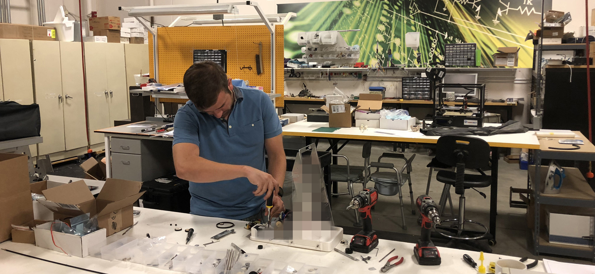 A man in a blue shirt works on assembling a device at a cluttered workbench in a workshop, surrounded by tools, small parts, and boxes. The background has workstations and a large mural with scientific imagery.