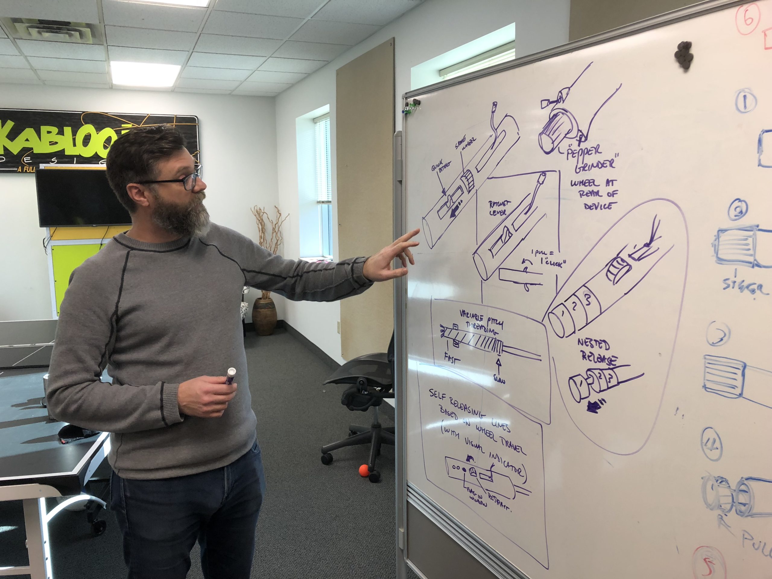 A man with glasses and a beard points at technical sketches and diagrams on a whiteboard in an office setting, holding a marker.