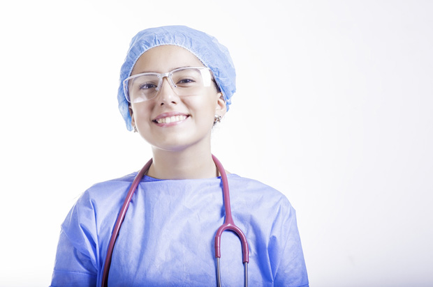 A smiling healthcare worker wearing a blue scrub, surgical cap, protective glasses, and a stethoscope around their neck, standing against a plain white background.