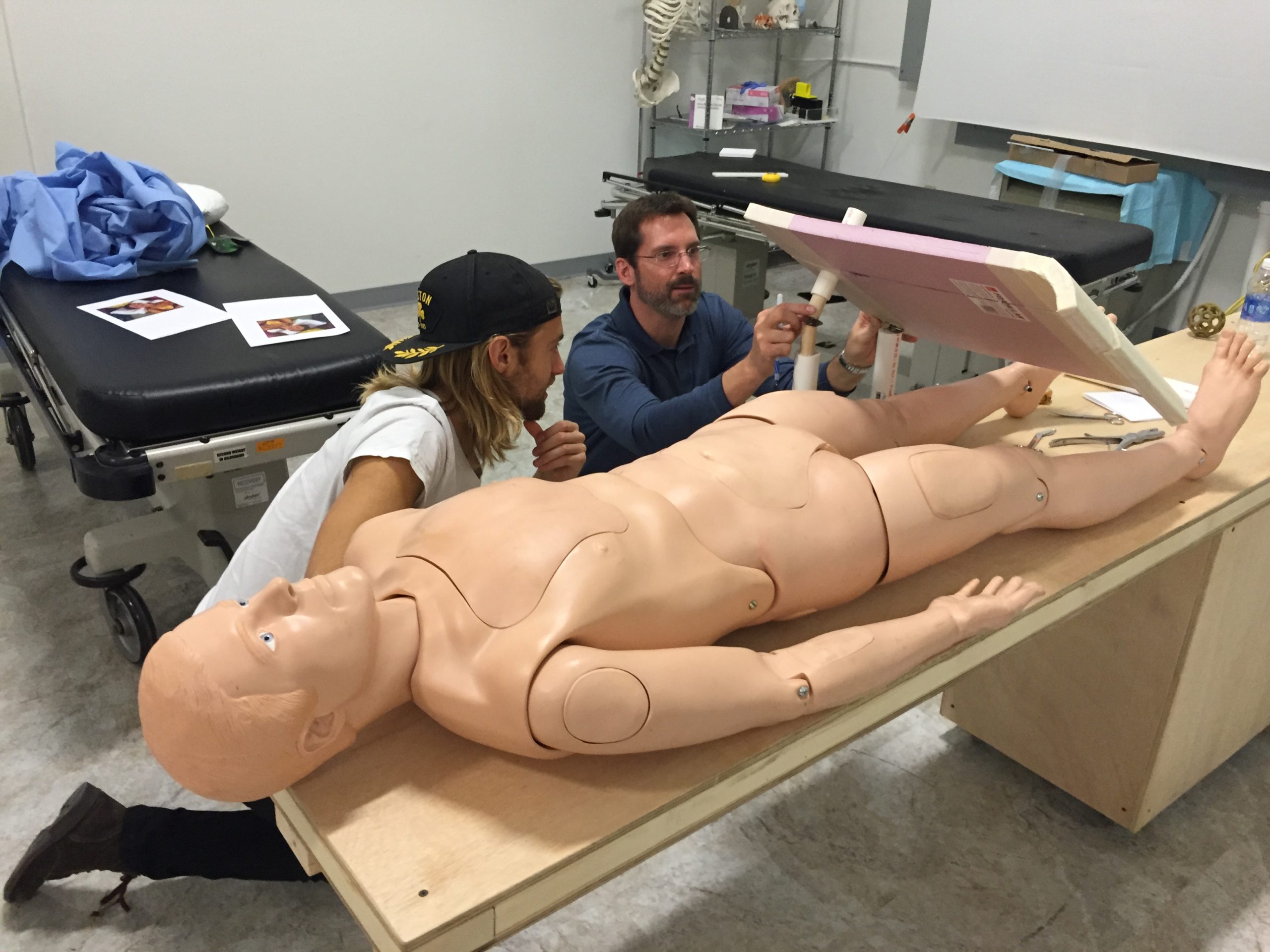 Two people kneel beside a medical mannequin lying on a table. They appear to be adjusting equipment or a panel near the mannequin’s upper legs in a clinical or training room setting.
