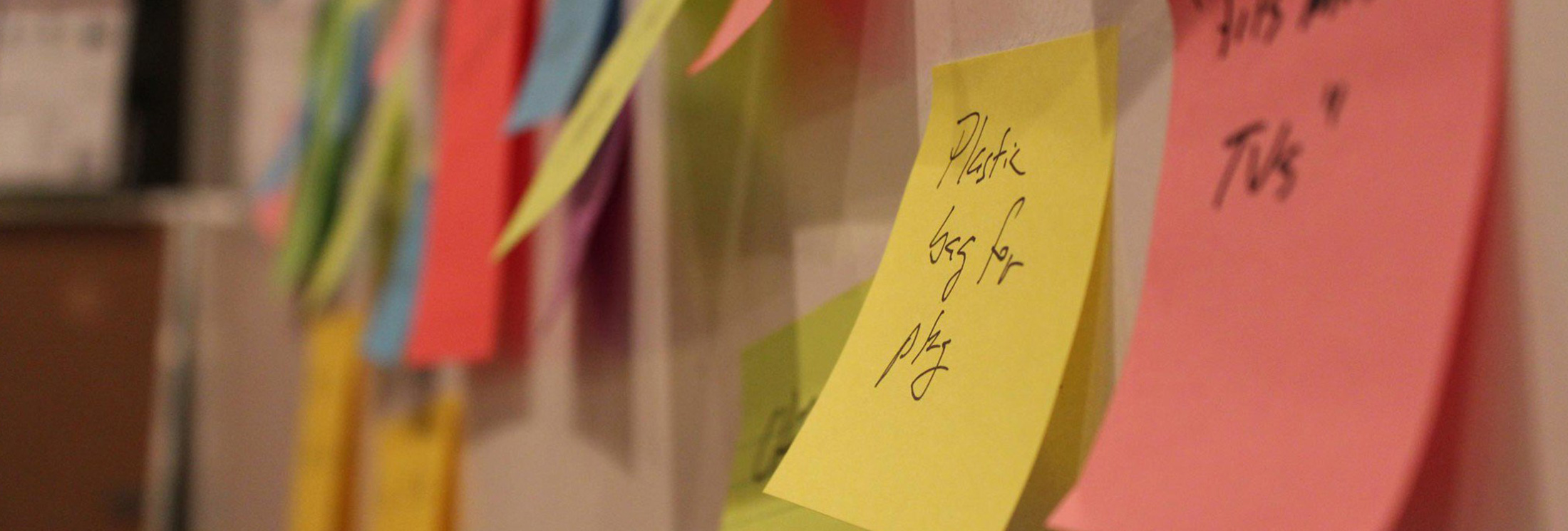 Close-up of colorful sticky notes attached to a wall, with handwritten text visible on some, including a yellow note that reads "Please beg for pity." The background is blurred.