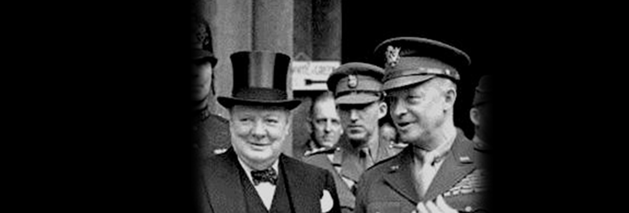 Black-and-white photo showing a man in a top hat and bow tie standing beside a uniformed military officer; both are smiling, with other people visible in the background.