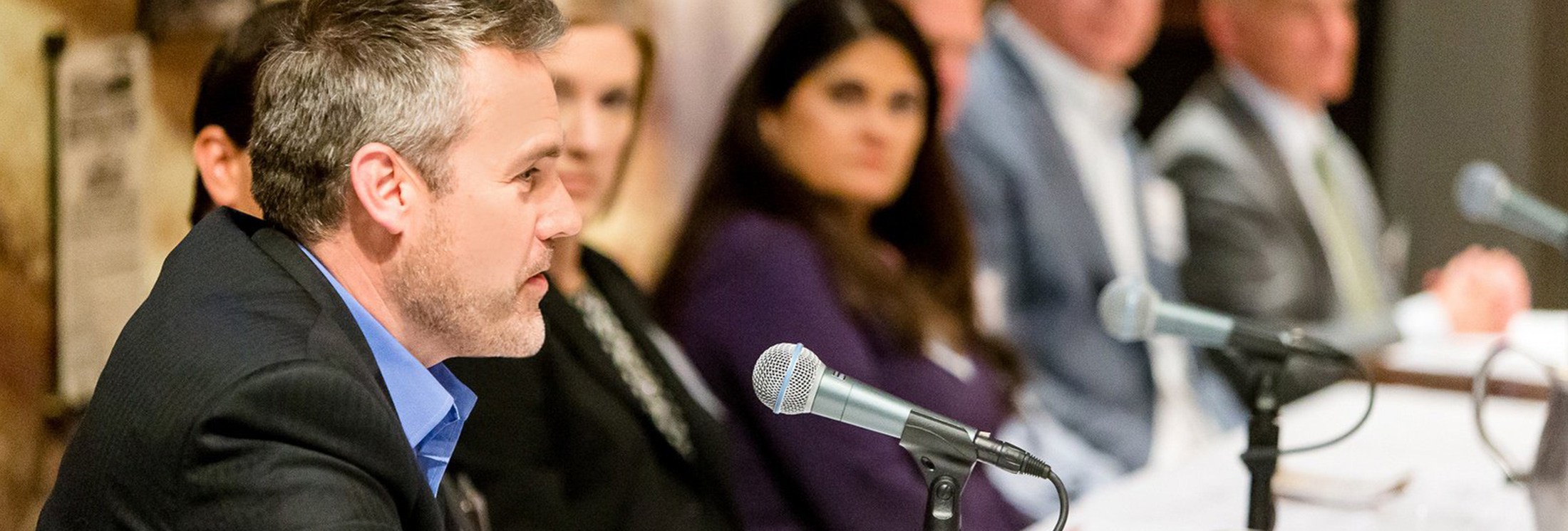 A man in a suit speaks into a microphone at a panel discussion, with several people seated in a row and listening in the background.