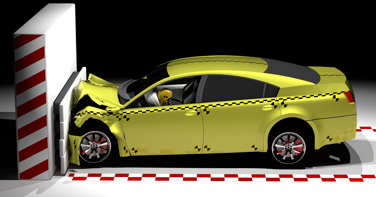 A yellow car with crash test markings is shown in a controlled collision, crashing head-on into a white barrier. The front of the car is crumpled, and a crash test dummy is visible in the driver's seat.