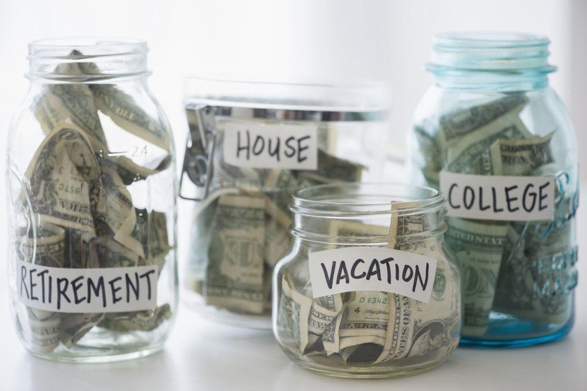Four glass jars labeled "Retirement," "House," "Vacation," and "College" are filled with assorted U.S. dollar bills, representing different savings goals.