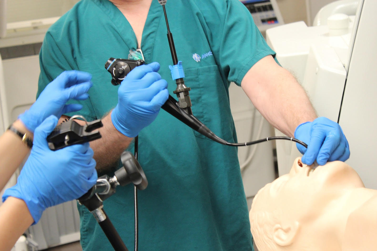 Two people wearing blue gloves use medical instruments to examine a mannequin's respiratory tract, simulating an endoscopy procedure in a clinical training setting.