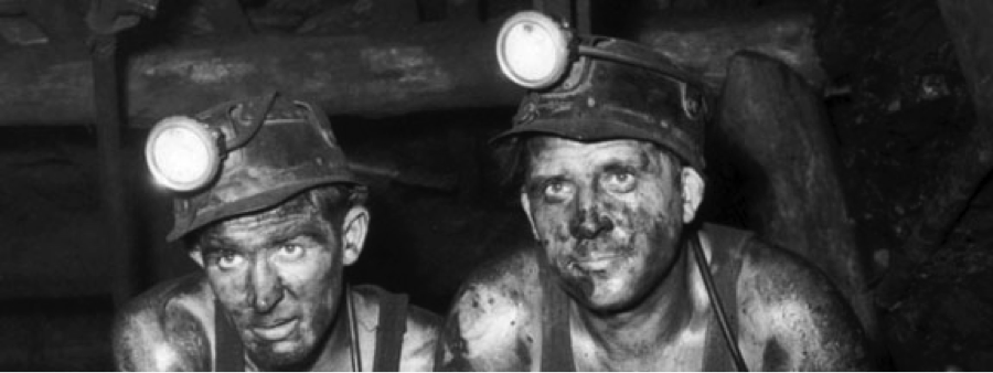 Two coal miners with dirt-covered faces and headlamps on their helmets sit side by side underground, appearing tired and focused, in a dimly lit mining tunnel.