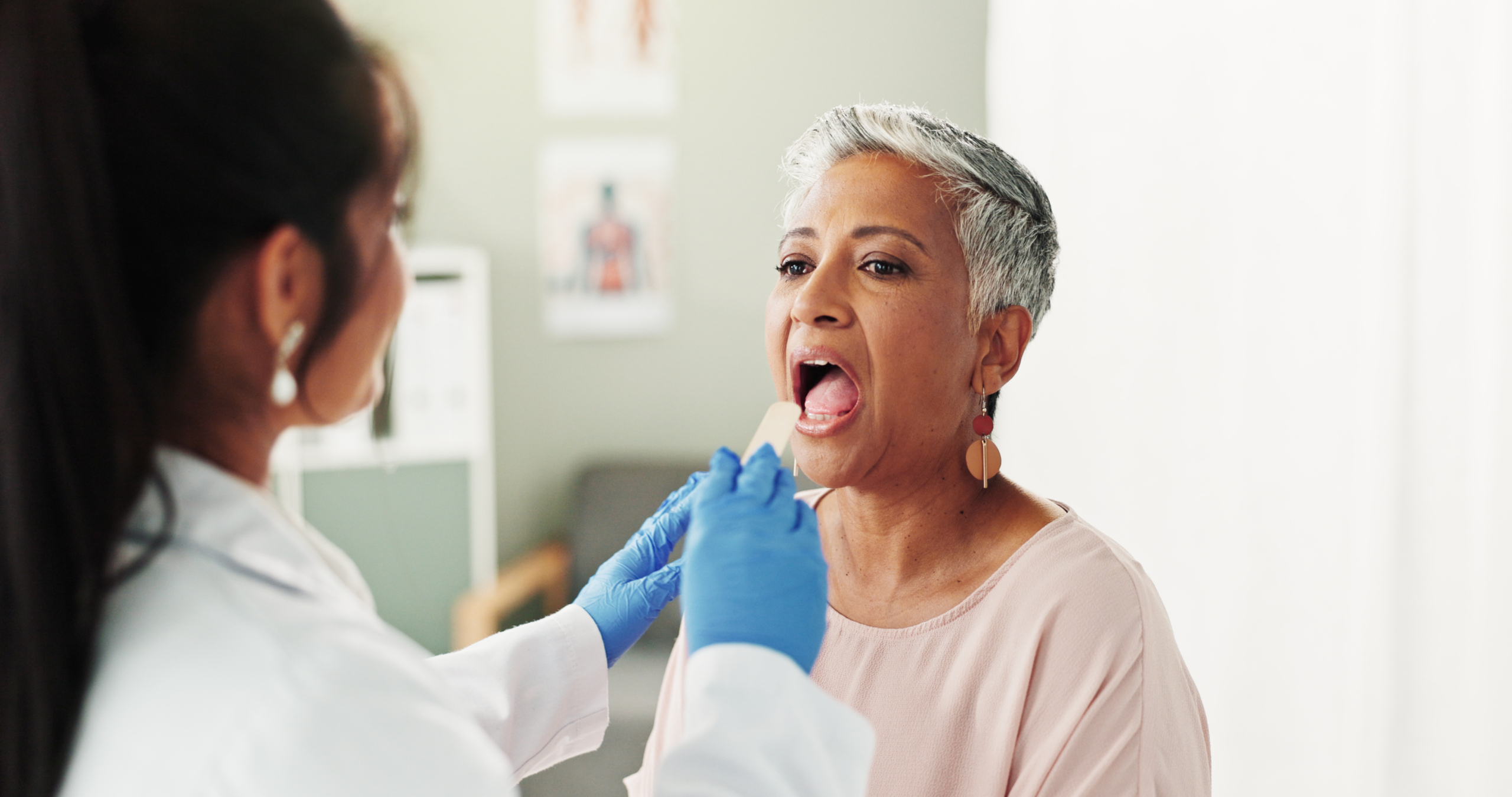 Clinic, woman and doctor with depressor for tongue, throat assessment and checkup for inflammation. Hospital, senior patient and otolaryngology with inspection for medical care, diagnosis and mouth